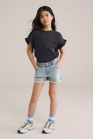 Fată în picioare cu mâinile în buzunare, purtând o bluză neagră cu mâneci volanate, pantaloni scurți din denim deschis, șosete albastre cu dungi și teniși argintii.