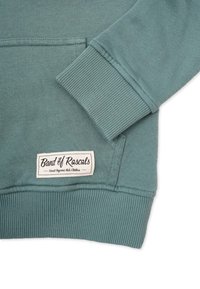 Teal-farbener Hoodie aus Biobaumwolle mit gerippten Bündchen und Saum. Verfügt über eine Fronttasche und ein Markenlabel, das am unteren Ende eingenäht ist.