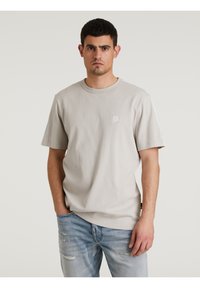 T-shirt in cotone grigio chiaro con scollatura a girocollo, maniche corte e un piccolo logo geometrico sul petto. Indossato con jeans azzurri scoloriti.