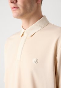 Ljus beige polo skjorta med en mjuk textur, med en kontrasterande krämfärgad krage och en rund, präglad logotyp på bröstet.