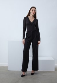 Bodysuit noir à manches longues avec un décolleté en V bordé de dentelle, associé à un pantalon noir à taille haute et jambes larges, et des chaussures à talons hauts noires.