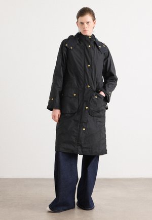 Zwarte waterdichte jas met gouden accenten, hoge kraag, voorzakken en knoopsluiting, gedragen over een oversized denimbroek.