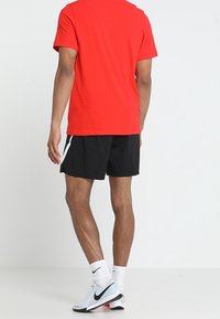 Mand står med ryggen til, iført en rød t-shirt, sorte træningsshorts med hvid stribe, hvide Nike-sokker og hvide Nike-sneakers.