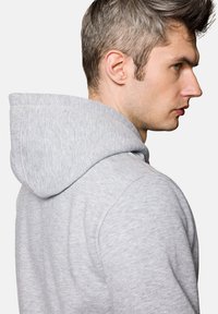 Szara bluza z kapturem i sznurkiem, o miękkiej fakturze, z podszewką wewnętrzną kaptura oraz widocznymi szwami. Widok z profilu użytkownika.