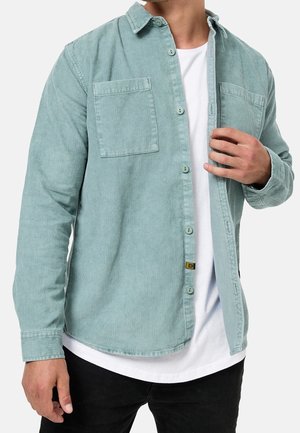 Man die een lichtgroen corduroy overhemd met knopen draagt over een wit t-shirt en zwarte broek, met één hand die de voorkant van het overhemd rechtzet.