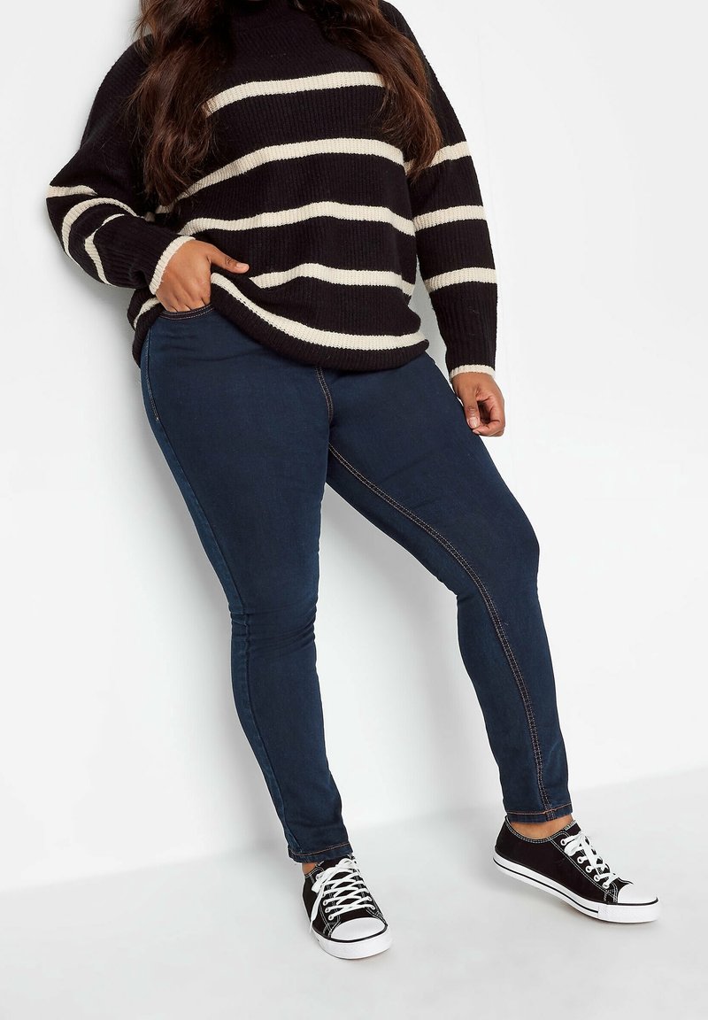 Pull en maille rayé noir et crème avec un col montant, associé à un jean slim bleu foncé et des baskets basses noires avec semelles blanches.