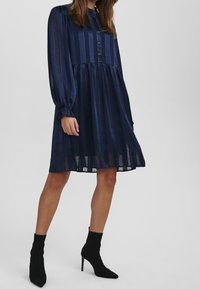 Granatowa sukienka w paski z długim rękawem, zapinana na guziki, z rozkloszowaną spódnicą. Przezroczysta tkanina, dopasowane mankiety i buty do kostek.