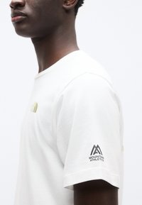 Λευκό βαμβακερό κοντομάνικο μπλουζάκι με λογότυπο σε ανοιχτό πράσινο χρώμα και μαύρο κείμενο «Mountain Athletics» στο δεξί μανίκι.