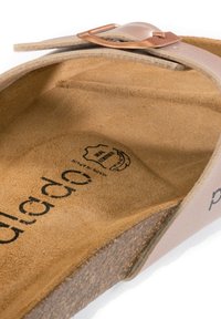 Sandalo in pelle con una cinghia rosa chiaro e una fibbia in rame. Suola interna in suede marrone morbido e suola in sughero. Dettaglio del logo impresso visibile.