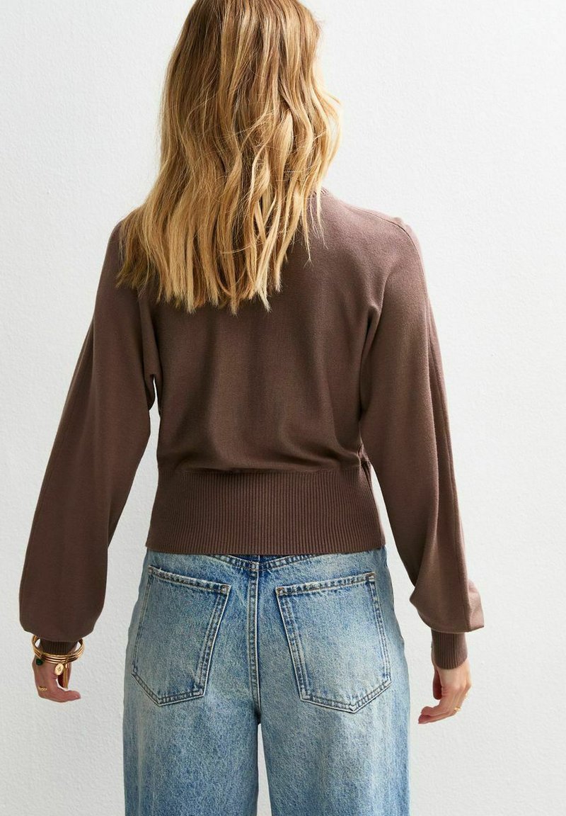 Eine blondhaarige Person, die einen braunen Langarmpullover und hochtaillierte helle Jeans trägt, steht vor einer weißen Wand.