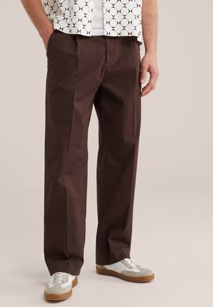 Férfi sötétbarna, egyenes szabású nadrágban, fehér tornacipővel és mintás rövid ujjú ingben, laza kéztartással áll.