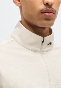 Κοντινό πλάνο ενός ατόμου που φοράει μπεζ ριγωτό τοπ με φερμουάρ μέχρι το στήθος, ψηλό γιακά και μικρό μαύρο λογότυπο στον γιακά.