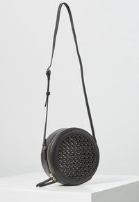 Borsa a tracolla rotonda nera con dettaglio intrecciato sul davanti, zip dorata e tracolla regolabile, esposta su un pedestal bianco.