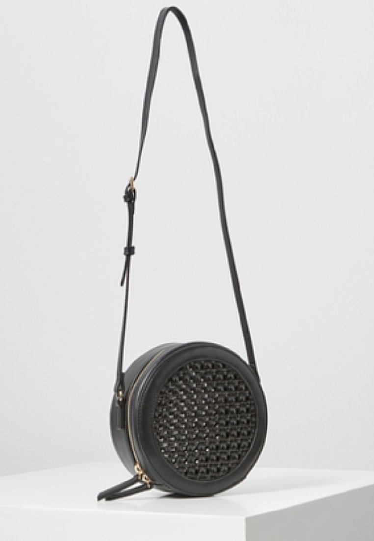Borsa a tracolla rotonda nera con dettaglio intrecciato sul davanti, zip dorata e tracolla regolabile, esposta su un pedestal bianco.