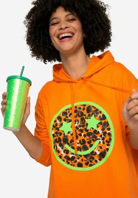 Felpa arancione con un ampio motivo di faccina sorridente a stampa leopardata contornato di verde. Il modello tiene in mano una tazza verde scintillante con una cannuccia.