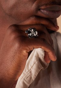 Silver lion head ring with intricate details and patterns, worn on a hand against a neutral background, showcasing textured skin.