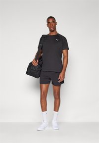 Schwarzes Sport-T-Shirt und Shorts mit einem Puma-Logo, kombiniert mit einer schwarzen Reisetasche. Weiße Sneakers und Socken runden das Outfit ab.