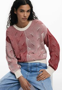 Rot- und cremefarbenes gemustertes Strickpullover mit Wellenmuster, runder Ausschnitt und gerippten Bündchen, getragen über hellblauen hochtaillierten Jeans.