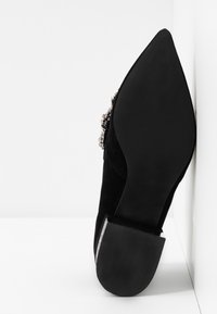 Chaussure à talon haut en daim noir avec un bout pointu, dotée d'un accent texturé en argent et d'un talon noir uni. Texture lisse, design épuré.