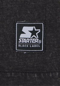 Fekete szövet, amelyen egy szőtt címke található egy fehér "S" és csillag mellett, a "STARTER BLACK LABEL" felirattal, kiemelve a letisztult vonalakat és a kontrasztos színeket.