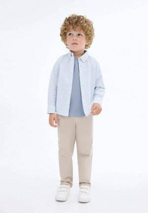 Cămașă butonată în dungi de culoare albastru deschis peste un tricou gri; pantaloni bej; adidași albi; păr creț; stând cu o expresie neutră.