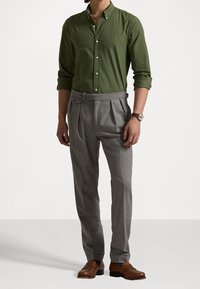 Groen overhemd met knopen en een klein logo, gecombineerd met grijze geplooide pantalon. Bruine leren schoenen maken de outfit compleet.