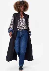 Long manteau noir sans manches sur un blouse rayé avec des accents bleus, associé à un jean bleu ample et des baskets noires avec des rayures blanches.