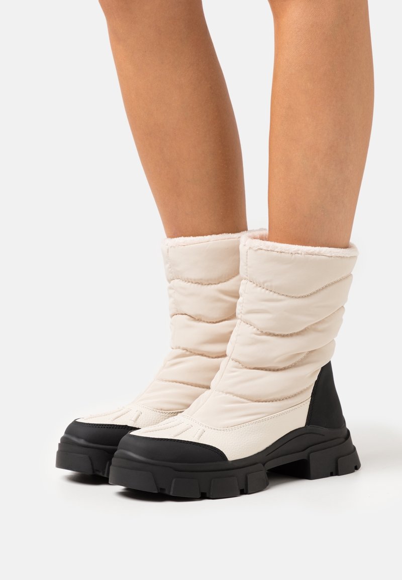 Jambes portant des bottes d’hiver rembourrées avec des semelles noires épaisses et des dessus matelassés beige clair atteignant le milieu du mollet sur un fond uni.