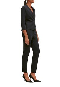 Vrouw in een strakke zwarte blazer met driekwartmouwen, bijpassende tapered broek en zwarte puntige hoge hakken, die zijwaarts staat.
