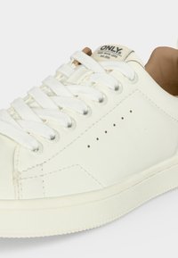 Close-up van een witte leren sneaker met witte veters, kleine ventilatiegaatjes en de tekst "ONLY Made with Love est. 1995" op het tonglabel.
