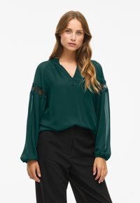 Blusa verde scuro con scollatura a V, dettagli in pizzo sulle maniche e maniche lunghe a palloncino semi-trasparenti. Abbinata a pantaloni neri.