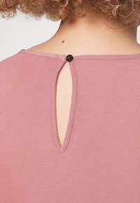 Rosa topp med en bakre nøkkelhull-design og en svart knappåpning, laget av teksturert stoff med en glatt finish.