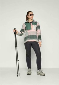Fleece-pullover i grønn, rosa og hvit geometrisk mønster med glidelås i kragen, kombinert med mørke bukser og grå tursko, og holder på turstaver.