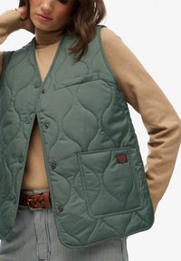 Kostkowany zielony kamizelka o teksturowanym wzorze, z kieszenią z przodu, zatrzaskami, noszona na beżowym swetrze, w połączeniu w paskowane spodnie.