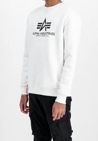 Sweat-shirt blanc à col rond avec un logo noir "ALPHA INDUSTRIES" et une étiquette sur l'ourlet, doté de poignets et d'un ourlet côtelés, fabriqué en tissu doux.