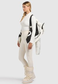 Biała bluzka z długim rękawem, dopasowane beżowe legginsy i kremowe botki za kostkę. Czarna i biała ocieplana kurtka zarzucona na ramiona, z widocznymi logo.