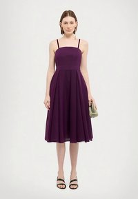 Vestido roxo, sem mangas, em linha A com pregas, corpete ajustado e alças finas; combinado com sandálias pretas e uma pequena mala verde.