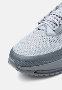 Ανοιχτό γκρι αθλητικό παπούτσι με επάνω μέρος από διχτυωτό υλικό, υφή και μαξιλαρ ωμένη σόλα με ορατή μονάδα αέρα. Σχοινάκια σε ταιριαστούς γκρι τόνους.