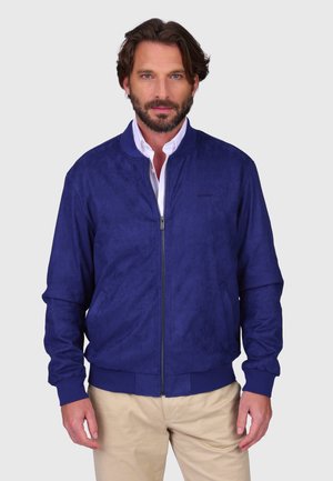 Homme barbu portant une veste zippée bleu marine sur une chemise blanche et un pantalon beige, faisant face à l'avant sur un fond uni.