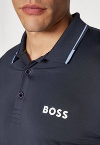Ναυτικό πόλο μπλούζα με κολάρο που κουμπώνει, με μπλε λεπτομέρειες και λευκό λογότυπο "BOSS" στο στήθος. Λείο ύφασμα με εφαρμοστή γραμμή.