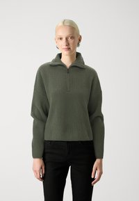 Olivgrüner gerippter Pullover mit hohem Kragen und halbem Reißverschluss. Verfügt über eine lockere Passform und heruntergezogene Schultern. Kombiniert mit schwarzen Hosen.