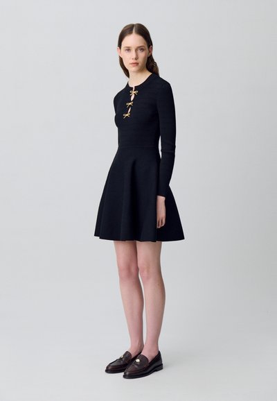 Donna che indossa un abito in maglia nero a maniche lunghe con tre bottoni decorativi a fiocco e mocassini marroni scuri, in piedi contro uno sfondo neutro.