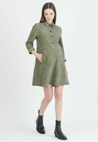 Abito in corduroy verde oliva con chiusura a bottoni, colletto da camicia, maniche lunghe e due tasche laterali. Forma a A con tessuto testurizzato.