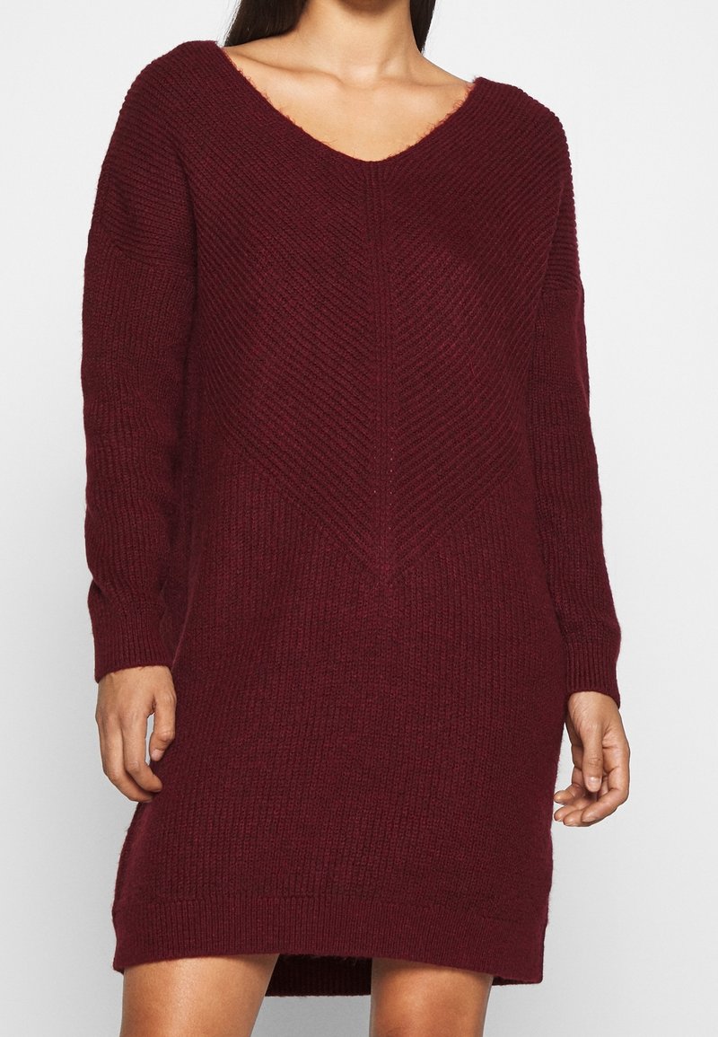 Robe tricotée bordeaux avec un décolleté en V, des manches longues et un motif texturé. Elle présente une coupe décontractée avec un ourlet droit et des fentes sur les côtés.
