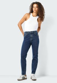 Visoko pasje temno modre kavbojke s klasičnim krojem, v kombinaciji z belim brezrokavnikom in belimi supergami z črnimi poudarki.