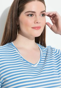 Modrý a bílý pruhovaný tričko s výstřihem do V, vyrobené z měkké tkaniny. Má volný střih a jemné detaily šití.