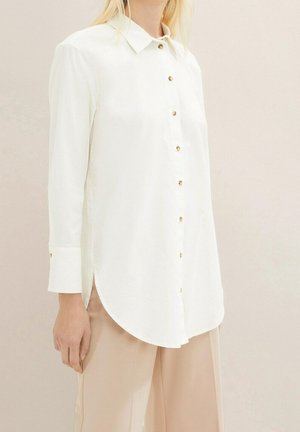 Vrouw draagt een witte blouse met lange mouwen met gouden knopen, in combinatie met beige broek, staand tegen een neutrale achtergrond.