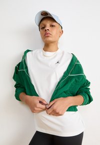 Grønn lett jakke med elastiske ermer, hvit t-skjorte med en liten svart logo, sammen med en lyseblå caps og svarte leggings.
