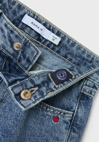 Pantalones de mezclilla con un lavado azul claro, que presentan botones de cobre y azul marino, una etiqueta blanca y un pequeño bordado de un corazón rojo en el bolsillo.