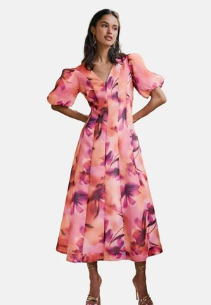 Kvinde iført en midikjole med puffede ærmer og pink-lilla blomsterprint, kombineret med gyldne sandaler med høje hæle og stropper.